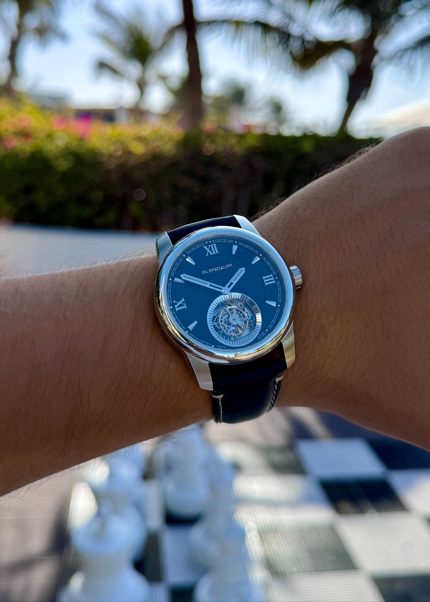 Person wearing a watch with a blue face and black strap, standing on a checkered floor with plants in the background.