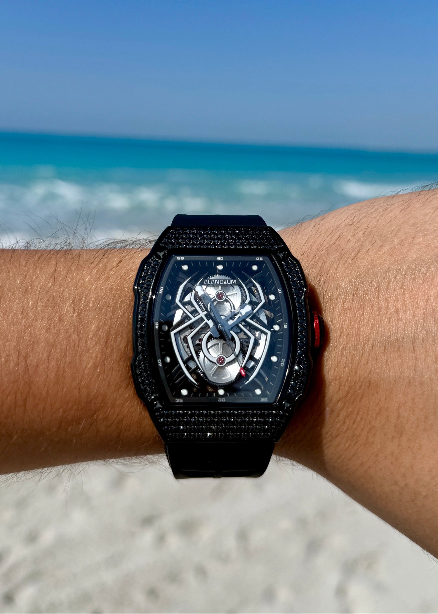 Black wristwatch with intricate design on a person's wrist against a beach background