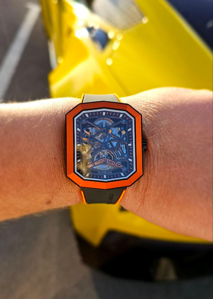 Watch with a blue face and orange frame on a wrist, with a yellow vehicle in the background.